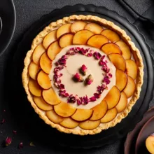 Plum custard tart decorated with sliced plums and dried rose petals on a black platter on a black table