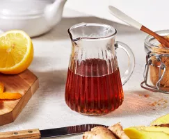 A small glass pitcher of golden brown sweet and spicy ginger syrup shown with sliced oranges and a jar of brown sugar.