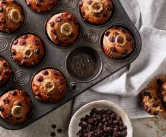 Vegan Banana Chocolate Chip Muffins in a muffin tin shown with one muffin removed and split open, and a bowl of chocolate chips.