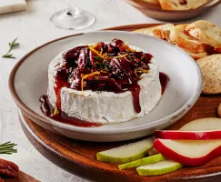 Baked brie topped with spiced candied pecans and Dark Brown sugar syrup and garnished with orange zest and rosemary, shown on a plate on a serving board with crackers, crostini, and sliced pears and apples.