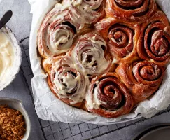 A round baking dish lined with parchment and full of cinnamon rolls, half of them iced, shown with a bowl of icing and a bowl of Dark Brown sugar