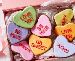 Heart-shaped sugar cookies decorated with royal icing with romantic messages written in icing, shown on white tiles with bowls of royal icing and a piping bag.