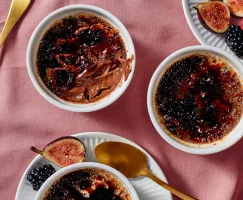 Three ramekins of salted dark chocolate crème brûlée, one partially eaten, shown with gold spoons, figs, and berries, on a rose-coloured tablecloth.