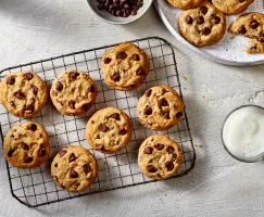 Chocolate chip cookies on a wire cooling rack with additional cookies on a platter, shown with a bowl of chocolate chips and a glass of white milk