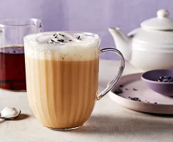  A glass mug of London Fog Latte with milk foam, shown with a pitcher of vanilla syrup, a tea pot, a stirring spoon, and a purple bowl of loose Earl Grey tea on a plate