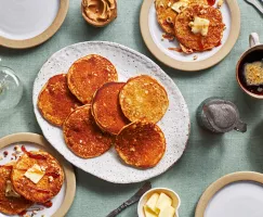 A table set with a platter of peanut butter pancakes, butter, a bowl of chopped peanuts, peanut butter, a mug of coffee, cutlery, a pitcher of syrup, and two plates with pancakes, syrup, and butter