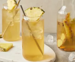 Two glasses of tepache with metal straws and garnished with pineapple wedges, shown with a pitcher of tepache