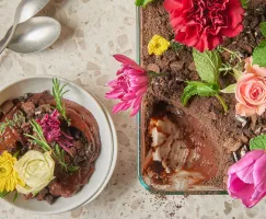 Chocolate dirt pudding with cookie crumble topping garnished with edible flowers in a glass baking dish shown with a serving of pudding in a bowl with spoons