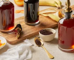 A bottle of coffee simple syrup, a bottle of orange simple syrup, and a bottle of banana simple syrup, shown on a table with a serving board, with coffee grounds, orange peels, and banana peels