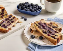Two slices of loaf pan French toast with blueberries drizzled with maple glaze and shown with a cup of coffee, a dish of blueberries, and the partially sliced loaf 