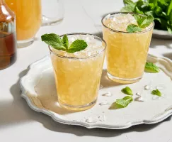 Two glasses of spirit-free mint julep with crushed ice and garnished with mint served on a white platter, shown with a bowl of mint leaves, half a lime, a pitcher of prepared julep and a pitcher of mint simple syrup