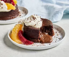 Two personal-size molten chocolate lava cakes on white plates, topped with whipped cream and cocoa powder, garnished with orange and blood orange segments, one shown with a piece missing and chocolate sauce spilling out from the centre.