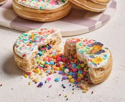 An iced pinata sugar cookie broken in two with candy confetti and sprinkles spilling out, shown with three more cookies on a white and mauve checkered tray in the background.