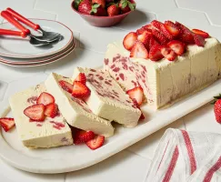 A rectangular loaf of strawberry-elderflower semifredo on a marble platter, garnished with sliced strawberries, with three slices cut, shown with three white plates with red trim, three red spoons, and a red bowl of strawberries.