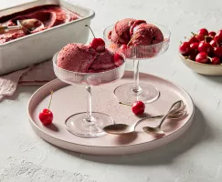 Two stemmed glass dishes of Cherry sorbet garnished with cherries, on a pale pink platter with gold spoons, shown with a bowl of cherries and a pan of cherry sorbet with a scoop for serving.