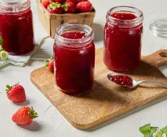 Three jars of strawberry jam, two on a wood cutting board and one on a tea towel, shown with a spoon of jam, fresh strawberries, and strawberry flowers.