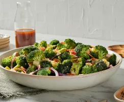 A serving bowl of broccoli salad with gochujang dressing on a kitchen counter shown with a pitcher of dressing, plates, and wooden serving spoons.