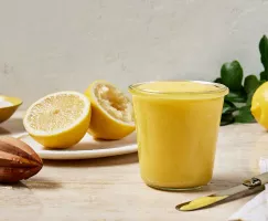  A glass jar full of lemon curd shown with a bisected lemon with one half juiced, a lemon juicer, and a bowl of granulated sugar.