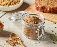 A lidded glass jar full of sweet Montreal steak seasoning shown with a raw, seasoned steak on a cutting board, a bowl of brown sugar, and a bowl of spices.