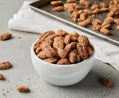 A white bowl overflowing with candied pecans and almonds, shown on a kitchen counter with a baking sheet of prepared nuts.