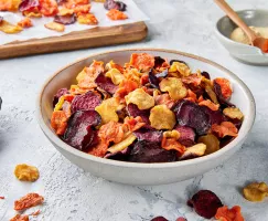 A stoneware bowl of mixed air fryer root vegetable chips shown with a bowl of Golden Yellow Sugar, a bowl of spices, and chips drying on a cutting board.