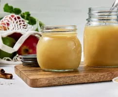 Two glass jars of homemade applesauce on a wooden cutting board on a white surface, shown with a mesh bag of apples, cinnamon sticks, and a bowl of Golden Yellow Sugar.