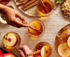 Four glass mugs of hot apple cider garnished with apple slices, cloves, and cinnamon sticks, shown on a table with a pitcher of cider, a bowl of apples, and a bowl of golden yellow sugar, and hands reaching for the mugs.