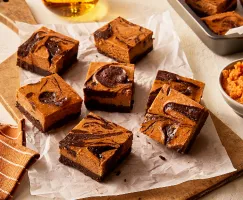 Seven pumpkin cheesecake brownies on a wooden cutting board lined with parchment, shown with a baking pan with brownies and a bowl of pumpkin puree.