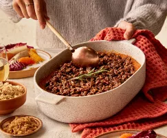 A woman wearing a sweater serving sweet potato casserole garnished with a rosemary sprig from a baking dish, shown with a radicchio and citrus salad, glasses of wine, and bowls of golden yellow and demerara sugar.