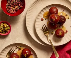 Balls of fried dough (Gulab Jamuns) with golden forks and small bowls filled with petals and pistachio