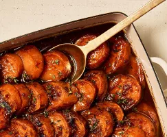 Sliced candied yams shown in a baking dish with a serving spoon, garnished with herbs and showing visible caramelization.