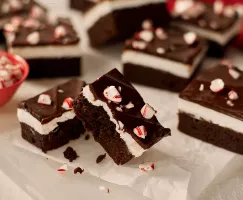 A close-up of chocolate mint brownies with a layer of mint cream and ganache top, each sprinkled with candy cane pieces, shown on wax paper on a white surface. 