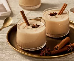 Two tumbler glasses of eggless eggnog garnished with anise, cinnamon, and cinnamon sticks, shown on a gold tray with anise and cinnamon sticks, and a gold stirring spoon beside