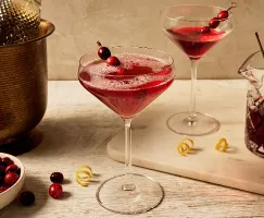 A stemmed glass filled with a carbonated cranberry cocktail, garnished with a skewer of cranberries and lemon zest, shown with a glass pitcher of cranberry cocktail, a stemmed glass of flat cranberry cocktail, lemon zest, and a bottle of sparkling wine in a brass ice bucket