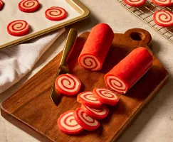  Two rolls of red and white peppermint pinwheel cookie dough on a wooden cutting board with several slices cut, with more slices on a baking sheet and baked cookies cooling on a wire rack.