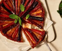 A carrot and parsnip tarte tatin resting on wax paper on a cutting board, shown with one slice partially removed with a pie lifter, garnished with fresh mint leaves.