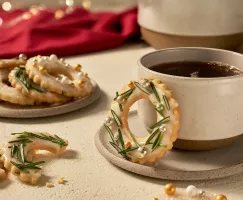 Glazed sugar cookies with icing and decorated like wreaths with rosemary, one on a saucer with a cup of coffee, one broken on a table, and several on a plate, shown in a festive holiday setting.