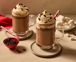Two glass mugs of vanilla hot chocolate topped with whipped cream and garnished with shaved chocolate and candy canes, resting on saucers and shown with marshmallows, shaved chocolate, and vanilla beans. 