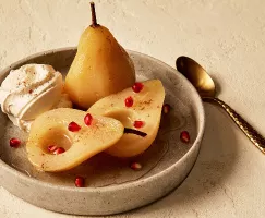 Three halves of white wine poached pears shown in a shallow dish, served with a dollop of whipped cream and sprinkled with cinnamon and pomegranate arils, and shown with a gold spoon.