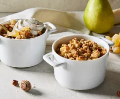 Two white single-serving bowls of pear ginger crumble, one with a dollop of whipped cream and a gold spoon, shown with a whole pear and a small dish of ginger pieces.