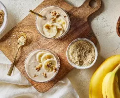  Top view of two tall glasses of banana oat tahini smoothie shown on a wooden cutting board with bowls of golden yellow sugar, sesame seeds, and dry oats with two unpeeled bananas.