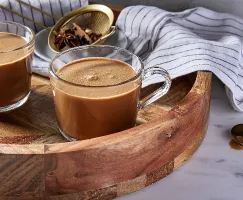 Two glass mugs of masala chai on a round wooden serving tray, shown with a gold strainer with whole spices in it, a dishcloth, and a gold spoon.