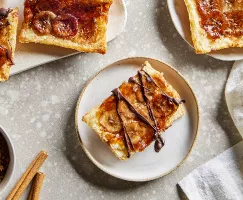 Four upside-down banana tarts, two on plates and two on a tray, shown with a bowl of brown sugar and cinnamon sticks.