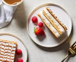 Two pieces of Mille-Feuille cake on plates, shown with berries, a cup of coffee and a gold fork.