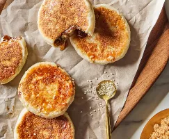 Five Korean pancakes or hotteok on a wood tray, two cut open with sesame syrup leaking out, shown with scattered sesame seeds and a bowl of Demerara Style Sugar.