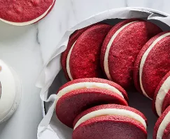 Red velvet sandwich cookies shown in a parchment-lined bowl beside a glass jar of milk with a paper straw.