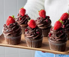 Six red wine chocolate cupcakes topped with ganache, shaved chocolate, pomegranate, and strawberries on a wooden tray held by a woman in a white sweater and jeans