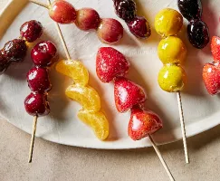 A platter of skewered fruit and berries coated in clear sugar syrup, or tanghulu, including mandarin segments, cherries, grapes, gooseberries, strawberries, kumquats, and blackberries