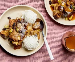 A serving of apple bread pudding on a plate with visible raisins and apple pieces, served with vanilla ice cream, shown with a second plate of bread pudding with butterscotch sauce and a glass pitcher of sauce.