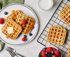  Three buttermilk waffles on a plate served with a pat of butter and strawberries and blueberries, shown on a kitchen counter with more waffles on a wire cooling rack, a glass of milk, and a bowl of mixed berries.